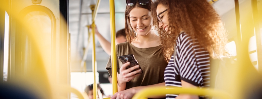 Two cheerful pretty young women are standing in a bus and looking at the phone and smiling while waiting for a bus to take them to their destination.