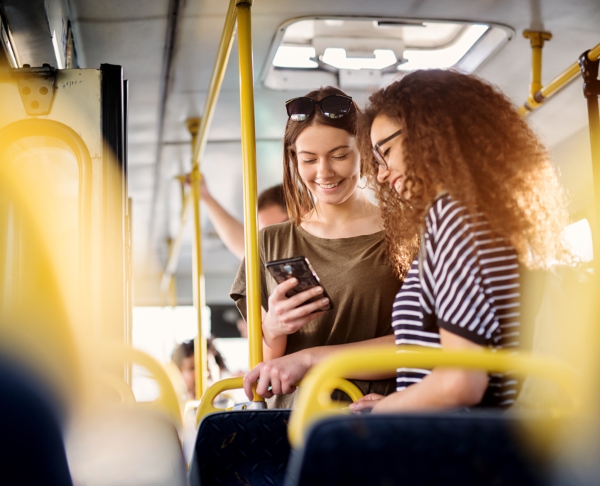 Two cheerful pretty young women are standing in a bus and looking at the phone and smiling while waiting for a bus to take them to their destination.