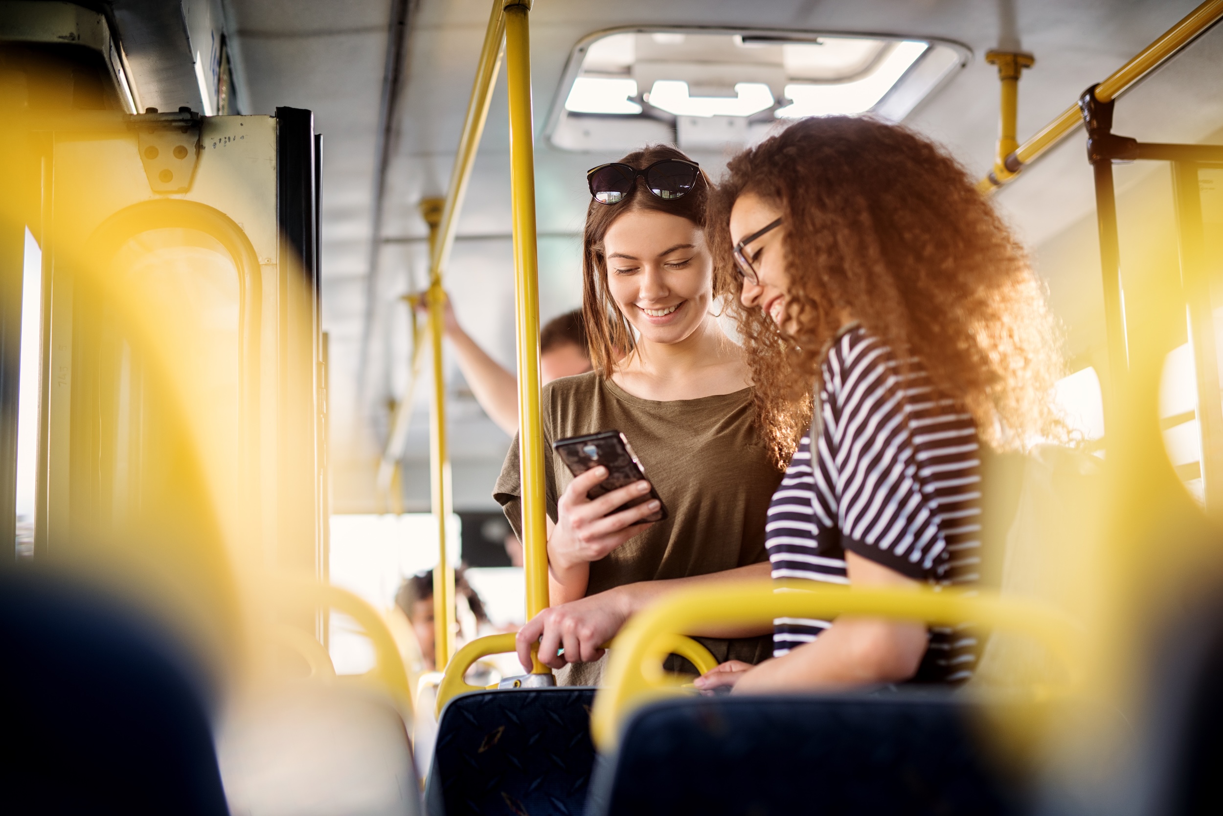 Two cheerful pretty young women are standing in a bus and looking at the phone and smiling while waiting for a bus to take them to their destination.